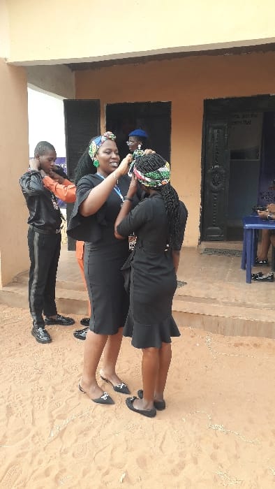 Two women stand embracing on a sandy surface, both wearing black dresses and traditional headwraps. One woman has her hand on the other's shoulder. A young boy wearing a tracksuit is visible in the foreground. They are outside a building with windows and doors, with a glimpse of people seated in the background. The scene appears to capture a moment of connection and celebration.
