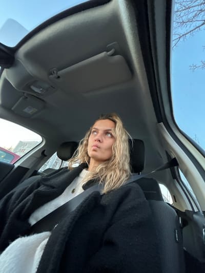 Inside a vehicle, a woman with curly blonde hair is angled towards the camera with a pursed-lip expression. She's wearing a white top and a black cardigan. The car's interior, including a sun visor and seat, is visible. The car window offers a glimpse of a building outside. The sky is visible through the front window. The seat belt is visible.
