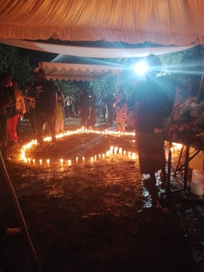 This image captures a nighttime outdoor gathering in Marenyo-Shianda ward, Kenya. The scene's primary illumination originates from numerous small, lit candles arranged on dark, wet ground, forming a heart-shaped outline. Overhead, a large, draped white fabric canopy provides coverage, beneath which a smaller white tent structure is visible further in the background. Multiple individuals are present, standing around the candle display. Most figures appear as silhouettes due to the low ambient light and an intense artificial light source flaring from the upper right-center, obscuring one individual. On the far right, a dark stand holds a floral arrangement, with a white bucket or container placed beneath it. Dark vegetation is visible in the background. The ground exhibits a wet, reflective surface. Individuals are observed standing, indicating participation in or observation of the event.