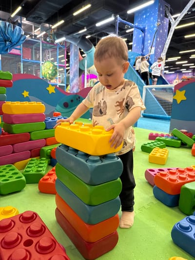 A young child is pictured playing intently in an indoor children's play center in Korobovo, Russia. The scene is brightly lit, likely daytime, and shows a vibrant, colorful environment.
The main subject is a young boy, with light brown hair, lying on his stomach on a lime-green soft play mat. He is wearing a light-colored t-shirt adorned with various animal prints, dark pants, and light-colored socks. His hands are engaged with large, oversized plastic building blocks, similar to Lego bricks but much bigger, stacking a yellow block onto a blue one. These colorful blocks, in shades of red, orange, yellow, green, blue, and pink, are scattered across the floor and form a tall structure next to him.
In the background, other children are visible within a multi-level blue climbing structure, which features mesh netting and star-shaped cutouts. One child in a white shirt with a red heart design and another in a light-colored top can be seen on a raised platform or bridge within the structure. The wider play area includes elements like a blue rabbit-shaped sculpture in the lower left and a glowing, spherical object (possibly a stylized planet or moon) hanging in the mid-ground. The ceiling is lined with bright fluorescent strip lighting. The overall impression is one of energetic and creative play in a safe, stimulating environment. No discernible text is clearly visible on any objects, though the red heart on a distant child's shirt is identifiable.