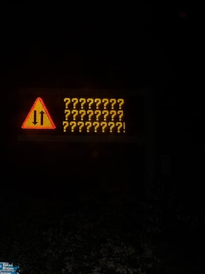 The image shows a roadside electronic sign in Cherniche, Bulgaria. The sign displays a yellow and red triangular warning symbol with arrows pointing in opposite directions horizontally. Above the symbol, the sign displays several lines of characters, possibly Cyrillic or Bulgarian, rendered as question marks, suggesting a malfunction or incomplete data. The surrounding environment is dark, indicating nighttime or low-light conditions. Trees are visible to the right of the sign. The background is generally dark.