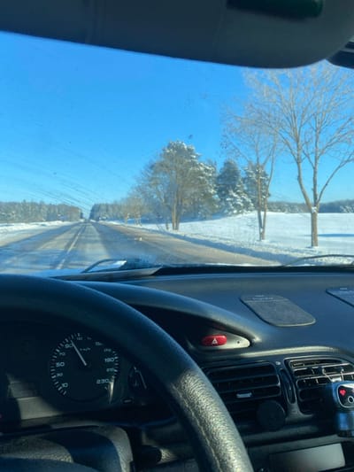 The image depicts a winter scene captured from the interior of a vehicle traveling on a two-lane road in Пліскі сельскі Савет, Belarus. The sky is clear and bright blue. The road ahead, which appears to be asphalt, is partially cleared of snow, revealing darker lanes, and is flanked by significant snow accumulation on both sides. Bare deciduous trees and snow-covered coniferous trees line the road and extend into snow-covered fields under a clear horizon.
Within the vehicle, a black, four-spoke steering wheel is visible on the left, obscuring the lower part of the instrument cluster. The speedometer, visible through the steering wheel, indicates a speed of approximately 90 km/h. The dashboard is dark grey, featuring a red, triangular hazard warning light button, two rectangular anti-slip mats, and air vents. A mobile device holder is clipped to the far-right air vent. The windshield exhibits streaks and minor debris, suggesting a recent cleaning or frost residue. No individuals are visible in the scene.