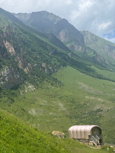 In the mountainous region of El'tiubiu, Russia, a covered wagon structure stands on a grassy slope. The wagon, with a light-colored fabric covering supported by a frame, features wooden wheels and a small set of stairs leading to an entrance. A person in a dark shirt is descending the stairs. The surrounding environment is dominated by lush green hills and the imposing, rocky peaks of the Caucasus Mountains, partially obscured by atmospheric haze and clouds. The foreground is a verdant meadow with scattered wildflowers.