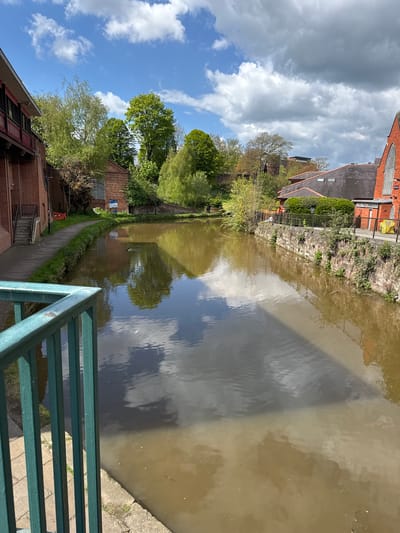 A canal scene, shot from an elevated perspective, possibly a bridge. The canal water is murky brown. Reflections of the sky, which features both blue patches and white, billowy clouds, are visible in the water. Buildings are on the canal banks, a mix of brick and other materials. Green foliage is present along the banks. A green metal railing is in the foreground. This is in Chester, United Kingdom.