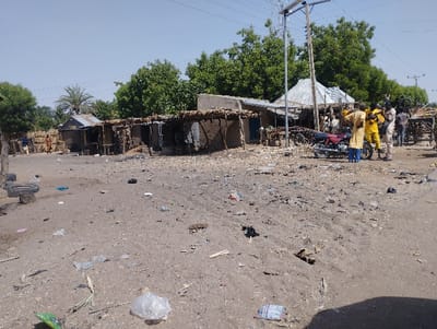 The image shows a dirt road in a small village in Kongo, Nigeria, located at latitude 9.726, longitude 11.148. The scene is littered with debris, including plastic bags, tire remnants, and pieces of wood. Six men are standing in the middle of the road, some are wearing yellow shirts and others wearing blue shirts. They appear to be talking to each other.  The image is taken from a slightly elevated perspective, looking down on the scene.  On the left side of the road there are a few buildings, including a small, single-story house with a corrugated metal roof, and some small shops with thatched roofs. There are trees behind the buildings. There is also a motorcycle parked in the foreground.  The sky is clear and blue, with no clouds. The road is dusty and dry.  The ground is covered in a layer of dirt and dust.