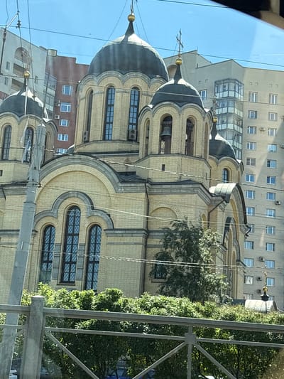 The image captures an outdoor daytime scene in Molodtsovo, Russia. The central focus is a large, pale yellow brick building, likely a church, with multiple dark domes topped with crosses. The building features arched windows with dark panes and decorative brickwork. To the left of the church, a tall, gray concrete utility pole is visible. Behind the church and to the right, several modern apartment buildings with numerous windows are present. Lush green trees and foliage are in the foreground, partially obscuring the lower section of the church. A metal fence runs horizontally across the bottom of the image. The sky is clear and blue.