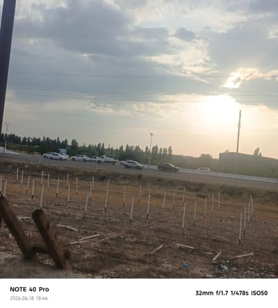 The image depicts a roadside scene in Sergeli, Uzbekistan, likely during the late afternoon or early evening, given the warm, diffused sunlight filtering through a cloudy sky. The foreground is a dry, barren field with numerous white posts planted in the ground, suggesting an area under construction or a vineyard. Two rough-hewn wooden posts are prominent in the lower left corner, appearing weathered and aged.

In the mid-ground, a multi-lane road stretches across the frame, with several vehicles, including white sedans, a white pickup truck, and at least one dark SUV, traveling in the same direction. The road is bordered by a low barrier and a dense line of trees forming a backdrop. Beyond the trees, some indistinct buildings and structures are visible, including what appears to be a tall, slender tower.

The sky is a dominant feature, filled with dramatic, layered clouds. Sunlight breaks through in the upper right portion of the frame, creating a bright, hazy glow. The overall impression is one of a rural or semi-rural setting on a somewhat overcast day.

Visible text in the image includes "NOTE 40 Pro" and "2026.06.18 18:46" in the bottom left, indicating the device used for capture and the date/time. In the bottom right, technical photographic information is displayed: "32mm f/1.7 1/478s ISO50".