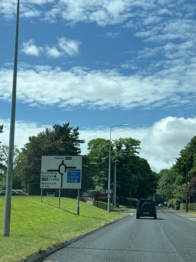 The image depicts a road scene on a sunny day, with a blue sky filled with scattered clouds. The focus is on a road sign indicating directions to various locations. The sign is predominantly white with black lettering. It shows directions to Chester (A 56), North Wales, Wrexham (A55/A483), Whitchurch (A 41), Ellesmere Port, Liverpool, and the M53. The sign is located beside a grassy verge and trees. There's a black SUV vehicle visible on the road in the background. Another sign is also visible indicating a "TOBY CARVERY 50".