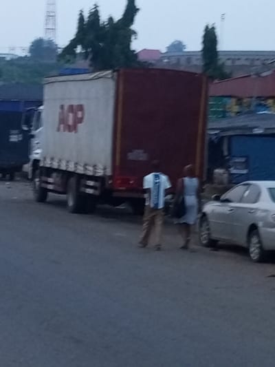 The image depicts a street scene in Oshogbo, Nigeria. A white truck with a red cargo box, bearing the red lettering "AOP" on its side, is parked. To the right of the truck, a silver sedan is visible. Two individuals are present: one person in a white and blue striped shirt and brown pants, and another person wearing a light blue dress and carrying a dark bag. The surroundings suggest an urban environment with various buildings and structures, including a water tower in the background. The lighting indicates it may be early morning or late evening.