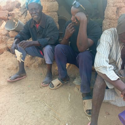 The image displays three adult males seated outdoors on a low, earthen structure adjacent to a dilapidated mud-brick wall in Belnin, Nigeria. The individual on the left, an elderly male, wears a blue patterned headwrap, dark long-sleeved shirt over a red undershirt, and light-colored trousers, holding a partially peeled, light-colored food item in his left hand. His gaze is directed forward. The central male, appearing middle-aged, wears dark sunglasses pushed onto his head, a black short-sleeved t-shirt, and dark blue trousers; his right hand is positioned near his mouth, and he looks downwards to his left. The third male, partially visible on the right, wears a light-colored striped long-sleeved garment and light-colored trousers, with his head also oriented downwards. All three men wear open-toed sandals. The ground beneath them is dry, light-brown dirt with scattered small sticks and debris. A broken white plastic bucket or container is visible on the ground behind the leftmost individual.