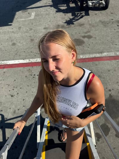 A young woman with blonde hair is standing on a metal staircase outdoors. She is wearing a white tank top with "LOS ANGELES CALIFORNIA" printed on it in black lettering, denim shorts, and flip-flops. She is holding a clutch purse in her left hand and reaching for the handrail with her right hand, which has red nail polish. The woman is looking down and smiling, with her eyes closed.
The background shows a gray, asphalt surface with some white and red markings. There are shadows cast by the staircase and some equipment, suggesting it is daytime and sunny. The scene appears to be at an outdoor location, possibly near an airport or a similar transportation hub, given the staircase and the industrial-looking equipment. The woman's expression suggests a happy or relaxed mood. There are no explicit indicators of the specific time of day, but the strong shadows suggest sunlight. The location is identified as Kruja, Albania, which might imply a tourist destination or a travel-related setting.