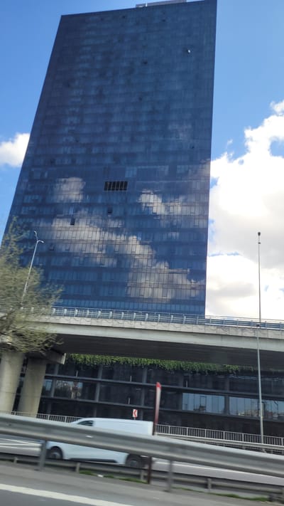 A tall, blue skyscraper with a mirrored facade dominates the left side of the frame, reflecting white clouds in a blue sky. To the right, a concrete overpass structure runs parallel to the building. Green foliage is visible on the side of the overpass closest to the skyscraper. A white van is partially visible in the foreground, partially obscured by a gray metal barrier. The scene is located in Bağcılar, Turkey. A streetlight is also visible.