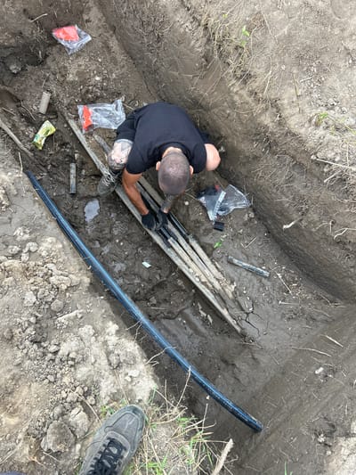A man wearing a black t-shirt and black gloves is kneeling in a muddy trench. He is working with several dark pipes laid out in the trench. There are various objects scattered around the trench, including red plastic cones in clear plastic bags, a cardboard tube, a yellow plastic bag, and other debris. A large black pipe with blue stripes runs along the bottom left of the frame. The ground is composed of dark soil with some dry grass and rocks. The image appears to be taken outdoors, possibly in a construction or utility work area. There is no specific indication of the city of Sadovo, Bulgaria, in the image itself.