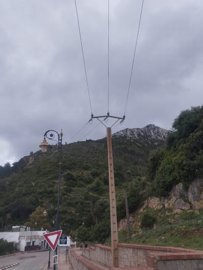 The image shows a street scene in Tétouan, Morocco, with a mountainous backdrop under an overcast sky. A red and white triangular yield sign stands near the road. A decorative lamppost is positioned beside the sign. A concrete utility pole with electrical lines extending upward dominates the center of the frame. A low brick wall runs along the side of the road. Vegetation covers the hillside, including trees and shrubs. Buildings are visible on the left side of the road. A rocky mountain rises in the background.