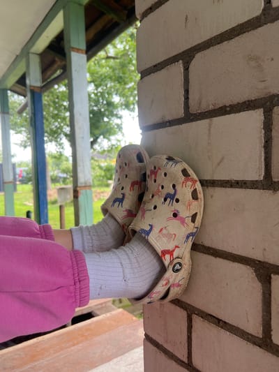 The image shows a close-up of a person's feet, wearing white Crocs with a pattern of pink and blue unicorns, and grey socks with holes, resting against a white brick wall. The person is wearing pink sweatpants. In the background, a wooden porch structure with painted blue, green, and light blue posts is visible, with trees and greenery beyond. The scene appears to be outdoors, possibly in a residential area.