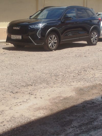A black Haval SUV is parked on a gravel surface in what appears to be an outdoor area. The vehicle is positioned at a slight angle, facing towards the left of the frame. The front of the car features a distinctive grille with the Haval logo. A license plate that reads "01 S 757 ZZ" is visible on the front bumper. The surrounding environment consists of rough, uneven gravel and some dirt. A wall or building is visible in the background behind the car. The lighting suggests it is daytime, with shadows cast on the ground, indicating a sunny environment.