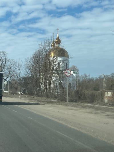 The image displays an outdoor scene in what appears to be late winter or early spring, with a prominent Russian Orthodox church as the central background element. The location is stated as Pokrovskoe, Russia.
In the foreground, an asphalt road occupies the lower left quadrant, displaying a faint white dashed centerline. To its right, a wide, unpaved dirt or gravel shoulder extends, indicating a roadside environment.
The main subject is a white church situated in the mid-ground, featuring at least two onion-shaped golden domes topped with Orthodox crosses. The church's white facade is partially obscured by numerous deciduous trees and dense brush, all bare of leaves.
To the right of the asphalt road, positioned between the road and the church, a circular regulatory road sign is visible on a grey pole. The sign displays a red circle with a diagonal line, depicting two cars (a red one on the left, a black one on the right), signifying "No Overtaking" or "No Passing."
Further to the right, utility poles with power lines are present. A rustic fence-like structure, heavily overgrown with dry vines or brush, defines a boundary in the right background. The sky is partially cloudy, with patches of blue visible amidst white, scattered clouds. No individuals or active interactions are discernible in the scene.