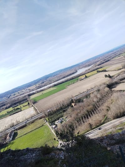 The image captures a vast, tilted aerial or high-vantage-point landscape view on a bright, clear day, likely late autumn or winter, given the bare deciduous trees. The perspective is looking down and across a wide valley or plain, with a slight diagonal tilt to the entire scene, making the horizon line appear sloped.
The top portion of the image is dominated by a pale blue sky, streaked with delicate, wispy white clouds that extend across the frame. Below the sky, the distant horizon reveals a low range of hills or mountains, appearing hazy and soft.
A prominent river or large body of water snakes its way through the mid-ground, reflecting the sky and appearing a light, silvery gray. It flows generally from the upper right towards the mid-left of the image. Along its banks and spreading throughout the valley are extensive areas of land segmented into various fields. Some fields are vibrant green, suggesting active agriculture or pastureland, while others are a lighter, muted beige or brown, indicative of fallow land, harvested crops, or fields awaiting planting.
Interspersed among the fields and along the river are numerous clusters and lines of trees. These trees are mostly bare of leaves, showing the skeletal structure of their branches, which further suggests a colder season. Some denser wooded areas form dark patches or strips across the landscape.
Human infrastructure is visible throughout the scene. A main paved road, with clear white lane markings, cuts diagonally from the bottom right quadrant of the image towards the mid-left, paralleling some of the fields and tree lines. Several vehicles, appearing as small dark shapes, can be seen traveling on this road. Another, narrower road or track, possibly unpaved, is visible further to the left, connecting different parts of the farmland. A railway line, distinguished by its straight, dark path, also runs through the landscape, generally parallel to the river and some of the fields.
Scattered buildings dot the landscape. A notable structure with a reddish-brown roof and light-colored walls stands near the bottom-left, adjacent to a green field and a road. Other smaller buildings or faint clusters of habitation can be observed in the distance, particularly along the riverbanks.
The immediate foreground, at the very bottom of the image, shows the rocky and somewhat rugged terrain from which the photograph was taken. This area is composed of rough, light-colored rocks and sparse, dark green foliage, including bushes and small trees. A faint, dark, wire-like fence structure is barely visible at the extreme bottom right corner, suggesting a barrier at the elevated viewpoint.
The overall impression is one of a vast, tranquil, and agricultural landscape, viewed from a considerable height under bright daylight. There is no visible text in the image.
