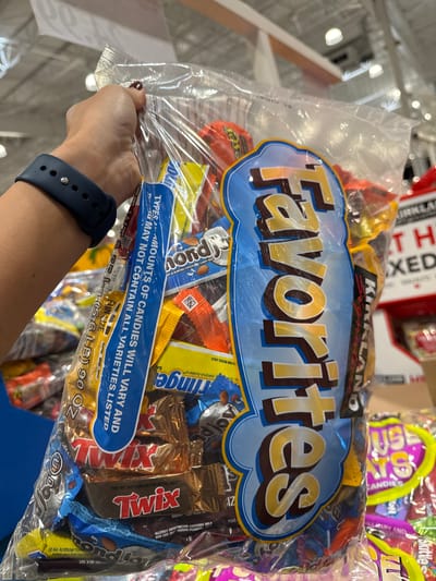 A person's hand, with dark red nail polish, is holding a large, clear plastic bag filled with assorted chocolates. The bag has a blue and yellow label that says "Favorites" in large, stylized letters. Individual candy wrappers are visible through the plastic, including Twix, Almond Joy, and what appears to be Reese's Peanut Butter Cups. A blue smartwatch is on the person's wrist. The background appears to be a retail store, possibly a warehouse club like Costco, with shelving and other products visible. The overall scene suggests a purchase of candy in Shoreline, Washington, USA.