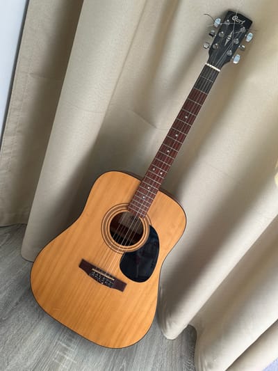 An acoustic guitar with a light wood finish and a black pickguard leans against draped, beige curtains in Zaboizkiy, Russia. The guitar is positioned diagonally, with its headstock towards the upper right and its body towards the lower left. The fretboard, made of a darker wood, features white fret markers. The headstock has six tuning pegs and the brand "Cort" is visible on the headstock. The guitar rests on a grey, wood-patterned floor. The curtains appear to be made of a textured fabric and are lit by natural light, casting shadows on the floor and the guitar. There are no people visible in the image.
