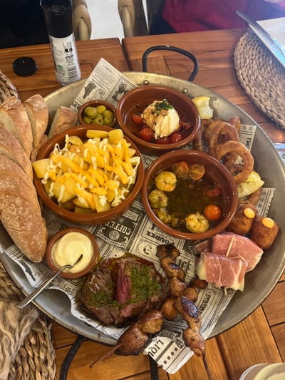 The image captures a large, round, metal platter brimming with various tapas. The platter is lined with newspaper-themed paper.
Food items include: onion rings, deep-fried croquettes with a yellow sauce, prosciutto-wrapped bites, a small bowl with shrimp in sauce, grilled chicken skewers, steak topped with green sauce, a bowl of fried potatoes with yellow and white sauce, olives, bread, and a small bowl of mozzarella and tomatoes. A small bowl of mayonnaise and a serving spoon are also visible.
A black bottle is lying on the table to the side of the tray. The table itself appears to be made of light-colored wood. A woven basket-like object is also present, likely serving as a place mat or decorative element.
The location appears to be a restaurant, presumably in Xàbia / Jávea, Spain, based on the traditional Spanish tapas served.