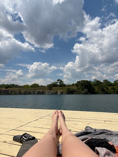 A person's legs and feet are visible, resting on a light-colored wooden dock. Beside them, a dark-colored slide with the text "GUESS" is present, along with some draped fabric, possibly towels or clothing. The dock is situated next to a body of calm, blue water. In the background, a tree-lined cliff face is visible, with rocky outcrops and dense green foliage. Above the cliff and water, a vast sky filled with dramatic, white and gray clouds stretches out, contrasting with patches of clear blue sky. The scene appears to be outdoors during the daytime, with bright sunlight casting shadows. The overall atmosphere suggests a relaxing moment by the water, possibly after swimming or sunbathing.