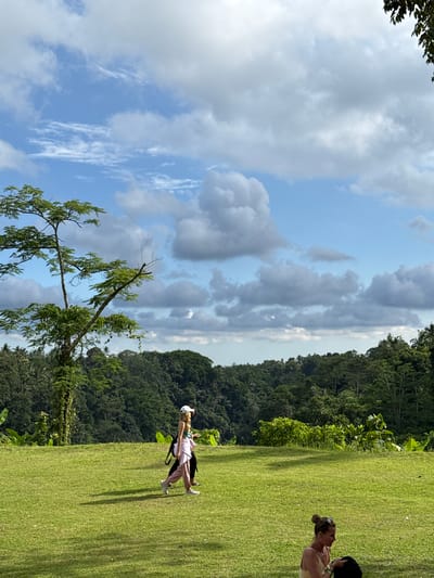 The photo shows a partly cloudy sky over a green grassy area near dense trees. Two women are present; one with dark hair is facing another, and the second woman is wearing a light-colored top and pants with a white hat, and a black bag on her back. The location is Carangsari, Indonesia. A tree with green leaves is present in the lower part of the photo.
