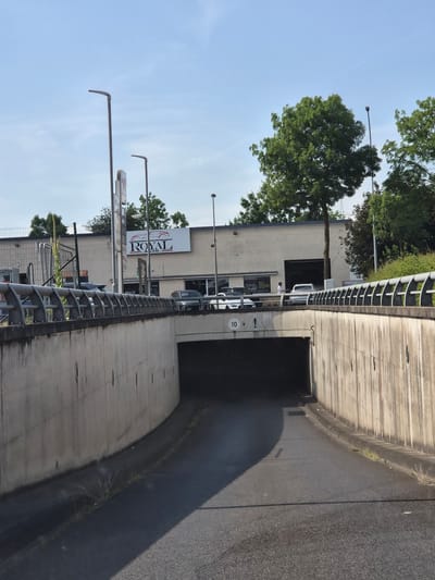 The image shows a tunnel entrance with a concrete structure on the right and a building with trees and a sign on the left. The sky is blue and clear, suggesting a sunny day. A shadow is cast across the tunnel floor, indicating it is likely daytime. The sign on the building reads "ROYAL AUTO". There are cars visible in front of the building and a person standing near the entrance of the tunnel. The overall scene appears to be an urban or suburban setting, possibly a car dealership or service center, with an adjacent road or pathway leading into a tunnel. The presence of streetlights and a speed limit sign (marked with "10") further supports this interpretation. The image is taken from a slightly elevated perspective, looking towards the tunnel entrance.