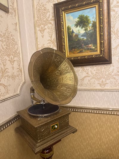 This media file displays a still life arrangement featuring an antique gramophone and a framed landscape painting. The gramophone is a prominent object, with its ornate brass horn and intricately decorated base. The horn is embellished with floral patterns and rests on a metal arm connected to the main unit. The base of the gramophone is a rectangular, metal structure, also adorned with decorative carvings and a small, framed image on its side depicting a white figure, possibly an animal, on a green background. A dark record is visible on the turntable.

To the left of the gramophone, a framed painting hangs on a wall with patterned wallpaper. The painting depicts a pastoral scene with a castle-like structure in the middle ground, surrounded by trees and rolling hills under a bright sky. Several figures and animals, including cows and a person with a horse, are present in the painting, suggesting a rural setting.

The background consists of textured wallpaper, with a light-colored floral pattern on the left and a warmer, more textured pattern on the right. The lighting appears to be ambient, suggesting an indoor setting. There is no visible text in the media file. The overall impression is one of a well-decorated interior, likely a home or a museum, showcasing vintage decorative items. The arrangement suggests a focus on historical artifacts and art.