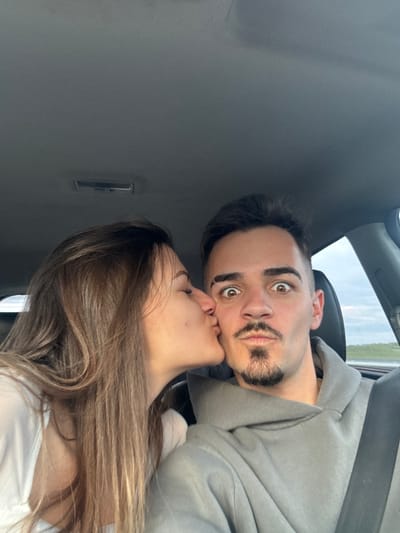 Two people are in a car. A woman with long brown hair is kissing a man. The man has dark hair, a beard, and is wearing a gray hoodie. They are inside a car with a gray interior. The location is unknown. The image does not provide sufficient information to determine if the scene is in the city of Chokoba, Bulgaria.