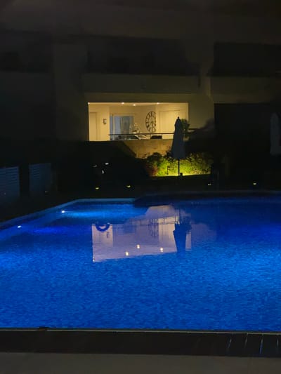 A night-time, eye-level outdoor photograph showcases a brightly lit, blue tiled swimming pool in the foreground. The pool's surface reflects the ambient light from underwater fixtures and the surrounding environment. In the background, a building is visible with illuminated windows and a large decorative clock on the exterior. An umbrella stands near a landscaped area with accent lighting. The overall scene suggests a resort or residential setting in Playa Del Sol-Villacana, Spain.
