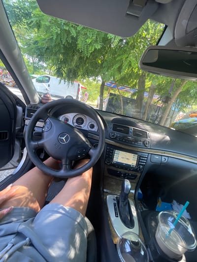 The image presents a detailed view from inside a Mercedes-Benz vehicle, capturing both the interior cabin and the external environment on a sunny day in General Todorov, Bulgaria.
The lower half of a person, identified by tanned legs wearing light grey shorts, occupies what appears to be the driver's seat. One hand, featuring long, light-colored acrylic nails, rests on the left thigh.
The car's interior features a black Mercedes-Benz steering wheel with integrated controls and an "SRS AIRBAG" emblem. The instrument cluster displays a speedometer reaching 260 km/h, and a digital readout indicates an external temperature of 29.5°C. A small, brown, spiral-shaped novelty item is positioned on the dashboard. The center console includes an infotainment system showing "107.7 MHz" and "FM," along with climate control dials. An automatic gear shifter with a silver-trimmed base is prominently featured. Wood-grain accents are visible on the dashboard and console surfaces. Two cup holders are present in the center console, one containing a clear plastic cup with a light brown beverage and a teal straw. The driver's side door is ajar.
Outside the vehicle, lush green trees with abundant foliage create a dense canopy. Through the trees and the open door, a white panel van is partially visible. Further in the background, informal market stalls or roadside shops, characterized by blue tarpaulin roofs and various displayed items, suggest a commercial or gathering area. Strong, direct sunlight illuminates the scene, creating distinct shadows and bright highlights.