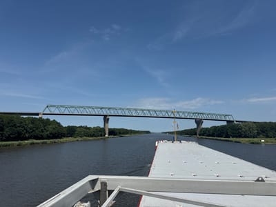 The image shows a view from a boat on a canal, looking towards a large green steel bridge. The canal is wide and dark, bordered by lush green trees on both sides. The sky is a clear, vibrant blue with a few wispy clouds. In the foreground, the deck of the boat is visible, painted in a light gray color with some red trim along the side. The bridge spans across the canal and appears to be a railway bridge. There are no people visible in the image. The overall scene depicts a calm, sunny day on the water, with the impressive structure of the bridge dominating the background. This scene is likely the Magdeburg Water Bridge in Brunsbüttel, Germany, which crosses over a canal.
