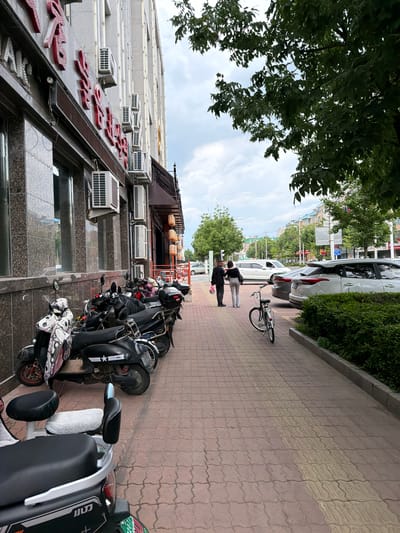 The image depicts a street scene in Hunchun, China, featuring a row of parked scooters and motorcycles along a sidewalk. A two-story building with air conditioning units and red Chinese characters on its facade lines the left side of the scene. The sidewalk is paved with interlocking bricks and has a tactile paving strip. Two individuals, a man and a woman, are walking away from the viewer on the sidewalk. A bicycle is parked nearby. Several cars are parked on the street beyond the sidewalk. Lush green trees and bushes are visible throughout the scene, and the sky is cloudy. The overall atmosphere is that of a typical urban street on a cloudy day.