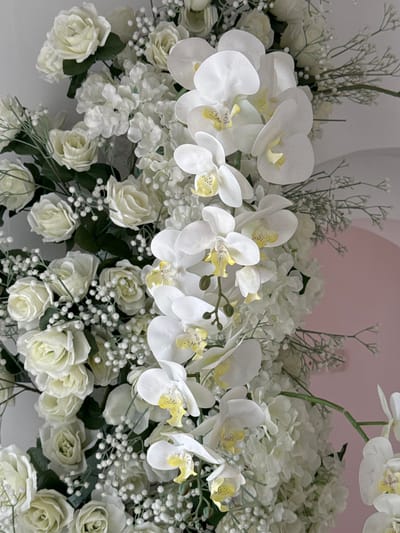 The image is a close-up, eye-level shot of an arrangement of artificial white flowers, primarily orchids and roses, interspersed with smaller white blooms and delicate green sprigs. The flowers are densely packed, creating a lush and voluminous display. The lighting is bright and even, highlighting the texture and detail of the petals. The background is a soft, out-of-focus gradient of pale gray and pink, suggesting an indoor setting with a curved wall. There are no people, animals, or any specific landmarks identifiable to pinpoint a location within Tanjungmeru, Indonesia. The scene is static and decorative, focusing solely on the floral composition.