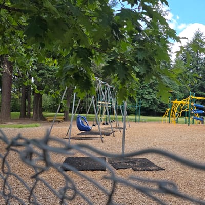 This image depicts an outdoor playground scene in Portland, Oregon, United States, viewed through a chain-link fence. The foreground is dominated by the blurred gray metal of the fence, which obscures a portion of the playground surface composed of wood chips and black rubber mats.

In the midground, a swing set with multiple swings is visible, with one blue swing prominently positioned. Behind the swing set, a yellow and green jungle gym structure is present. Further back, a yellow picnic table is situated near several large trees with thick trunks. The background consists of dense green trees and a bright blue sky with white clouds. The overall environment appears to be a park or recreational area. No people are visible in the image.