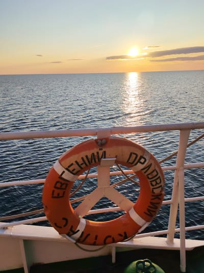 An orange lifebuoy hangs on a white railing aboard a vessel, facing a calm, dark blue sea under a pale sky with a setting sun. The lifebuoy has white stripes and black Cyrillic lettering that reads "ЕВГЕНИЙ ДРАВДСКИЙ" (Yevgeny Dravdsky). The sun, low on the horizon, casts a shimmering reflection on the water. In the foreground, a green object with a black valve is visible on the deck. The scene is set at the Kakkarovo location, Russia.
