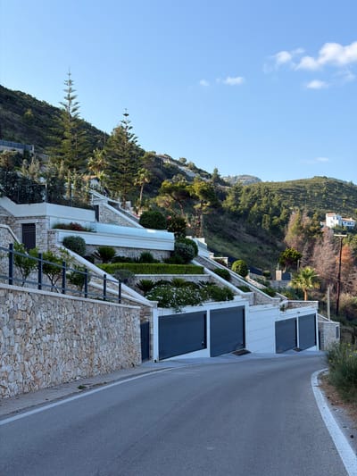 The image captures a view in the Municipal Unit of Sfakiates, Greece. The setting is a hillside location with modern architecture integrated into the landscape. A road runs along the right side of the image, adjacent to structures with stone retaining walls and white-trimmed buildings featuring dark garage doors. Lush greenery and trees are interspersed with the buildings, creating a terraced effect. The sky above is a clear blue with scattered clouds.