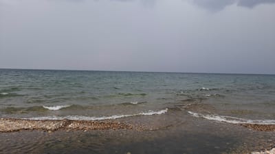 This is a photograph of a body of water under an overcast sky. The water appears to be a lake or sea, with gentle waves lapping at a pebble beach in the foreground. The sky is a uniform gray, suggesting cloudy or perhaps stormy weather. The horizon line is clearly defined, separating the water from the sky. The pebble beach is composed of small, smooth stones in various shades of brown and gray. The water is relatively clear, allowing the submerged pebbles to be seen in the shallows. There are no people or animals visible in the image, nor any man-made structures. The overall impression is one of natural, serene, and somewhat somber landscape. This scene is consistent with the general environment of Priboi, Russia, a coastal town known for its natural beauty and its location on the coast of the Baltic Sea.