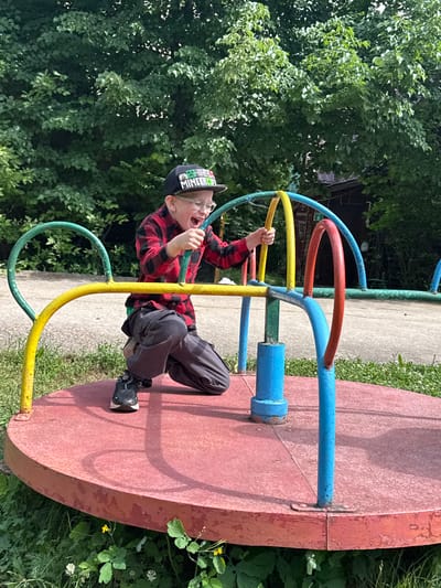 In Yunost', Russia, a young boy is on a playground merry-go-round. He is wearing a red and black checkered shirt, dark pants, and a black baseball cap with a "Minecraft" logo. He is leaning forward, holding onto the merry-go-round's colorful bars, with his mouth open as if shouting or laughing. The merry-go-round has a red platform and a frame painted in yellow, green, blue, and red. The background consists of lush green trees and a paved area.