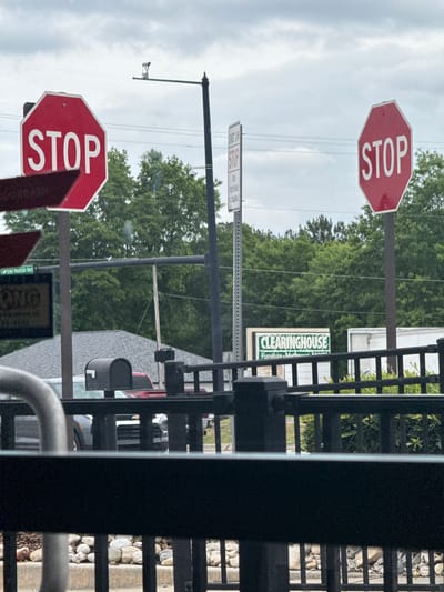 The image shows a scene in Stone Mountain, Georgia, United States. There are two stop signs and a sign that says "Clearinghouse Furniture, Mattress, and More". A street sign for Stone Mountain Hwy is visible. There is a black metal fence in the foreground. The weather is overcast. There are trees in the background. A utility pole and wires are visible. Cars are parked on the street.