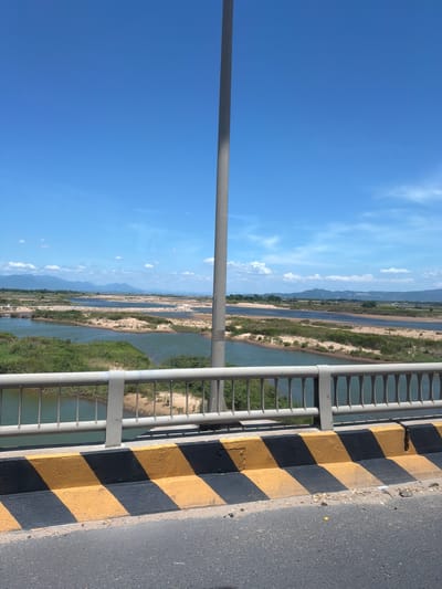 The image shows a view from a bridge, looking out over a river and its surrounding landscape. The scene is dominated by a vast expanse of bright blue sky with scattered white clouds. A tall, grey metal pole extends vertically on the left side of the frame, likely a lamppost or part of the bridge's structure.

To the right of the pole, the river flows through a green, vegetated valley. The river itself appears wide and shallow in places, with sandy banks visible. Distant mountains, shrouded in a light haze, form the horizon. The overall impression of the landscape is one of natural beauty and a rural setting.

In the foreground, the edge of the bridge is visible. A grey metal railing runs along the side of the bridge, providing a barrier. Adjacent to the railing is a concrete barrier painted with alternating black and yellow stripes, a common visual cue for hazard warnings or road boundaries. The road surface itself, a grey asphalt, is also visible on the far right of the image.

The bright sunlight and clear blue sky suggest it is daytime, likely a warm and sunny day. There are no people visible in the image, and the scene appears peaceful and serene. The location is identified as Tuy Hoa, Vietnam, which aligns with the lush greenery and mountainous backdrop. The image captures a scenic view of a river valley from a bridge, highlighting the contrast between the man-made structure and the natural environment.