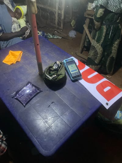 The image shows a close-up view of a blue plastic table with various objects on it. In the foreground, a person wearing a blue garment and a yellow hat is partially visible, seemingly asleep. Beside them, on the table, are a folded orange cloth and a sealed plastic pouch filled with a purple liquid. Further on the table, a rolled-up patterned fabric is placed next to a smartphone. The phone's screen displays a login prompt. A banner with large red letters on a white background is positioned over the table, with part of the word "DU" visible. The scene appears to be outdoors, possibly at night, given the lighting. The setting suggests a casual, perhaps rural environment in Sauka, Nigeria. There are no clear indications of weather, but the lighting suggests it is dark. The overall impression is one of a quiet, possibly resting moment.