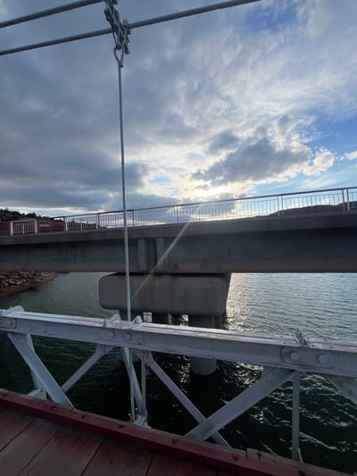 The image shows a view under a bridge in Ait Mazigh, Morocco. The bridge is constructed of concrete and steel with a gray color. Below the bridge is a body of water, with sunlight reflecting off its surface. In the foreground, a gray steel support structure of a different bridge or walkway is visible. The sky is cloudy, with patches of blue visible through the clouds.