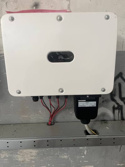 A Huawei solar inverter is mounted on a concrete wall in what appears to be an industrial or utility space. The inverter is white with a black Huawei logo in the center. Below the inverter, a black junction box is attached, with wires extending downwards. Red and black wires are seen entering the junction box from the top, and a white and yellow cable exits from the bottom. The wall has a rough, grey texture and is marked with some graffiti. A metal conduit or tray runs horizontally below the inverter and junction box, with the white and yellow cable feeding into it. The scene is illuminated by artificial light. The exact location within the city of Rozzano, Italy, is not discernible from the image itself.