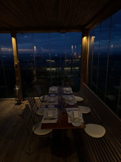 A long dining table is set for a meal in a building with large glass windows overlooking an evening scene. The table is covered with plates, silverware, glasses, and napkins. Surrounding the table are white chairs. The interior of the building has a wooden aesthetic with textured walls and plank floors. The view through the windows shows a dark sky and a distant cityscape or shoreline with scattered lights.