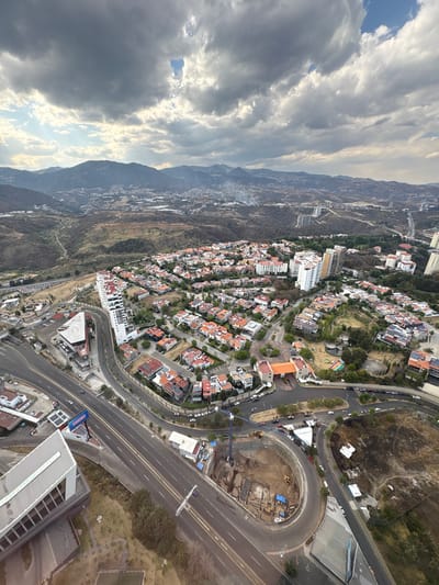 This high-altitude aerial image depicts an expansive urban and semi-rural landscape within Santiago Yancuitlalpan, Mexico, under a partially cloudy sky with breaks of sunlight. The foreground features a complex intersection of multi-lane asphalt roads, curving significantly. Adjacent to the main road on the right is a large, active construction site with a deep excavation pit, visible earthworks, multiple yellow construction vehicles (likely excavators), and a tall blue crane positioned centrally. Blue tarpaulins cover portions of the excavated area. A modern, multi-story building with a distinctive angular, white facade is prominent in the lower left, with another commercial-style building featuring a red-orange roof nearby.
The midground is dominated by a densely developed residential area, characterized by numerous houses with orange or reddish-brown tile roofs, interspersed with multi-story white apartment buildings. Roads snake through this development, and some roofs display solar panels. A distinct gated community entrance with a central roundabout, flanked by a tan-colored building, is visible, leading to an organized residential layout with green spaces and a small park containing colorful playground equipment.
The background transitions into a vast, rugged, and mountainous terrain, heavily vegetated with sparse urban development stretching into the distance. Distant hills are covered in trees and scattered structures, indicating further urban sprawl and rural areas. No individual people are discernible, but numerous vehicles are present on the roads, indicating active traffic flow. The overall scene portrays a dynamic urban environment undergoing expansion and development, set against a natural, hilly backdrop.
