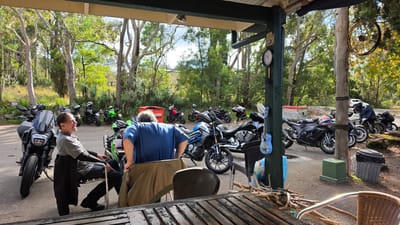 The image captures a sunny outdoor scene at a roadside establishment in Mount White, Australia, likely a cafe or biker-friendly stop, situated within a wooded environment. In the foreground, a wooden table with visible wood grain and metal railings suggests an outdoor seating area. Two men are present. One man, seated on a metal chair with a black jacket draped over it, faces left and appears to be laughing. He is wearing a grey long-sleeved shirt and dark trousers. The second man, wearing a blue t-shirt, stands with his back mostly to the camera, facing a row of motorcycles.
A collection of motorcycles is parked behind the men. From left to right, there is a large, dark motorcycle with a prominent windscreen, followed by several other motorcycles of various colors, including green, black, and a blue and grey model. Some of these motorcycles have helmets on them, and a blue decorative guitar is hanging on a green pole near the foreground.
In the background, a vibrant green wooded area with tall trees dominates the landscape. There are glimpses of other structures, including a red bin with "REMUNDIS" written on it, and what appears to be a clock on a wooden post. The lighting indicates a bright, daytime setting. The overall atmosphere suggests a gathering of motorcycle enthusiasts taking a break in a natural, semi-rural location.