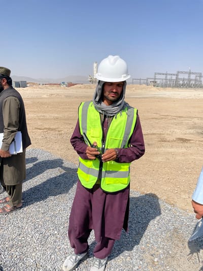 An outdoor image depicts three males on a construction or industrial site in Pir Dust, Afghanistan, under a clear blue sky. The foreground features light-colored gravel. A male in the center wears a white hard hat over a gray-and-white patterned headscarf, a fluorescent yellow safety vest with reflective strips, a maroon tunic, matching trousers, and light-colored sneakers. He holds a black handheld radio in both hands, looking downwards. To his left, another male stands, partially visible from the waist up, wearing a dark cap, a dark green tunic, dark trousers, and sandals. He holds a stack of white papers in his left hand. A third male's lower right arm and light blue clothing are visible on the far right. The background consists of vast, dry, light-brown earth. In the mid-ground, various structures are visible, including several shipping containers, a tall white industrial plant (possibly concrete), and a large electrical substation with multiple metal lattice towers. Distant hills are faintly visible on the horizon. Long shadows are cast on the gravel in the foreground, indicating bright sunlight.