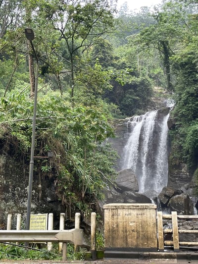 The image captures a waterfall cascading amidst lush greenery in Ramboda, Sri Lanka. The waterfall has three distinct tiers, plunging over rocky formations. A concrete structure and metal guardrail are visible on the right side of the frame, suggesting a roadside observation point. A lamppost stands in the foreground. Vegetation, including trees and shrubs, covers the surrounding landscape. The atmosphere appears humid or misty.